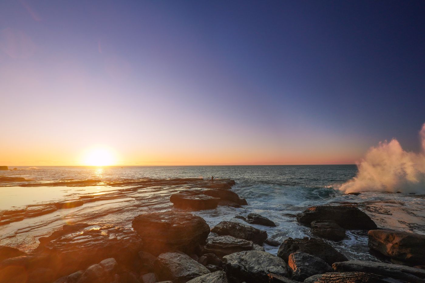 Sunrise over the rock platform with a wave exploding beside a lone angler
